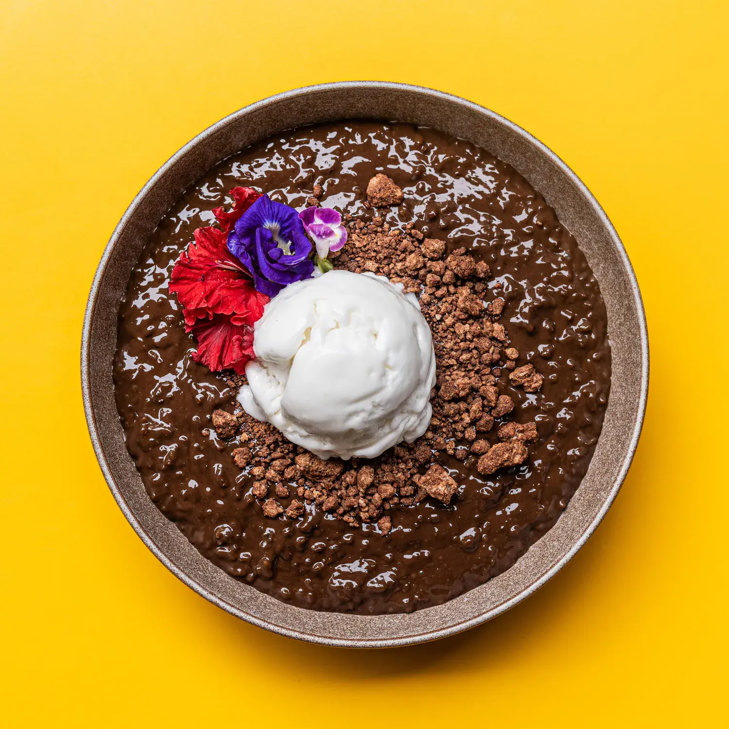 Champorado in a stoneware bowl with coconut ice cream, chocolate soil and edible flowers, shot from above on yellow